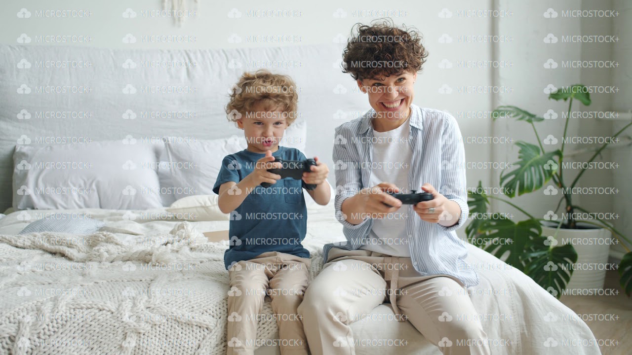 Mother and son playing video game sitting on bed in bedroom at home having fun